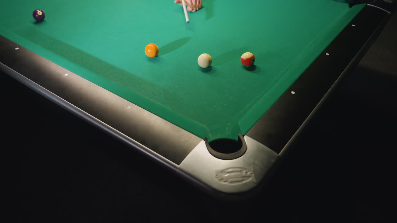 Up shot of hand using cue stick to strike white billiard ball on green pool table. Focus on scattered billiard balls and pocket, highlighting precision, movement, and tension in strategic gameplay