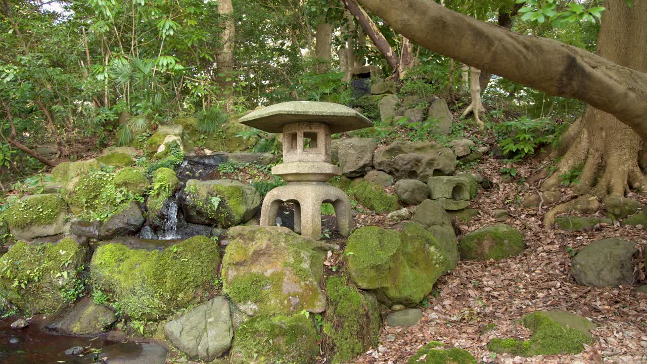 Inquadratura ampia e statica di una lanterna di pietra accanto a una piccola cascata in un giardino lussureggiante
