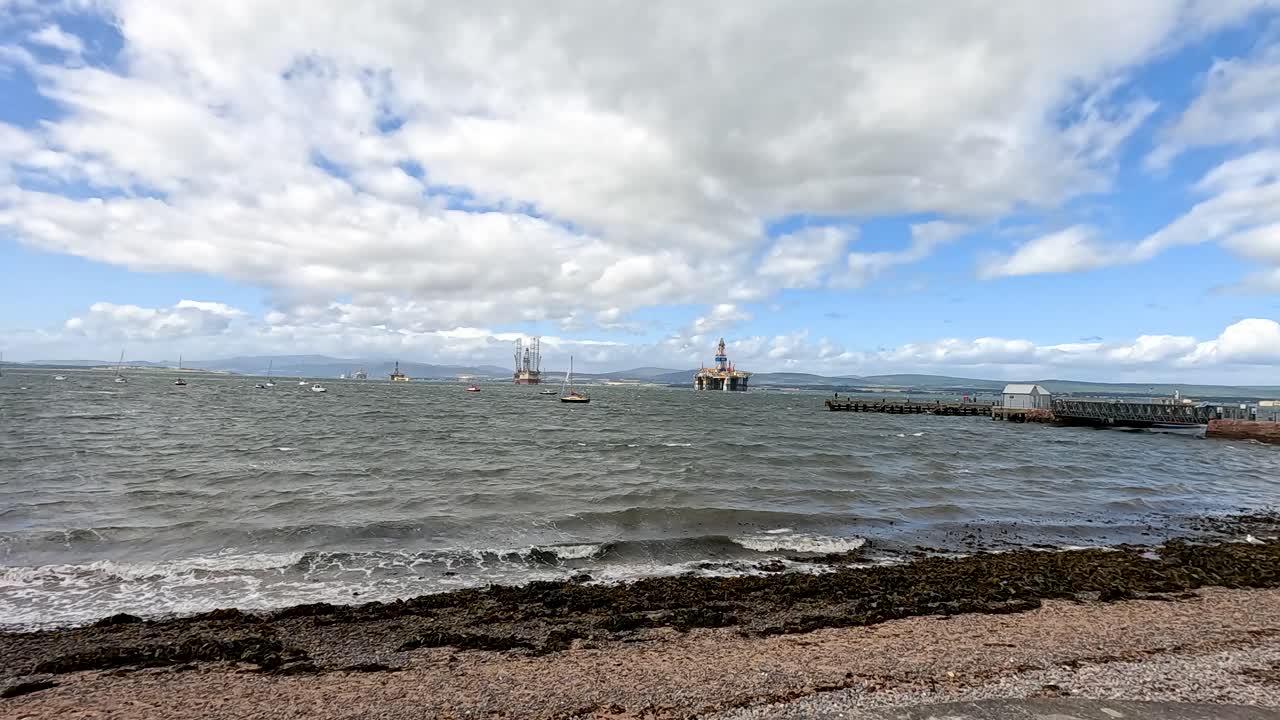 Waves roll onto a rocky Cromarty beach as sailboats and fishing boats drift offshore. Cloudy sky, dynamic lighting, and slow panning camera movement