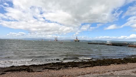 waves roll onto a rocky cromarty beach as sailboats and fishing boats drift offshore. cloudy sky, dynamic lighting, and slow panning camera movement