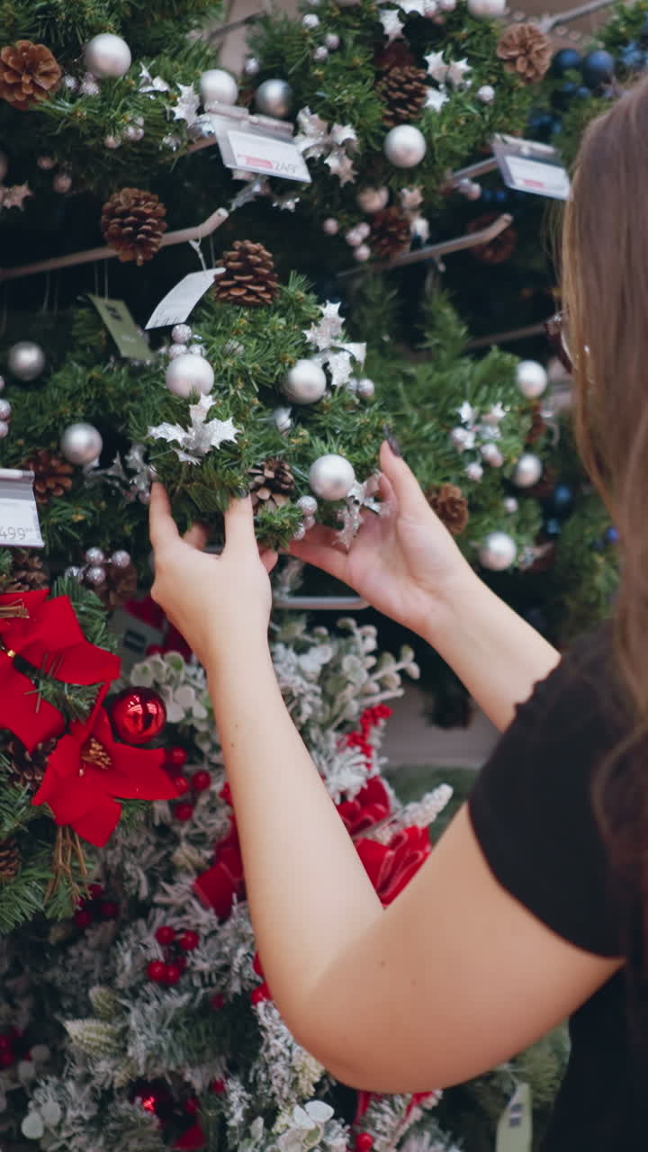 mujer joven con gafas y cabello marrón largo en camisa negra navegando por coronas festivas decoradas con adornos y piñas en la sección de vacaciones de la tienda con luces brillantes y señal direccional roja