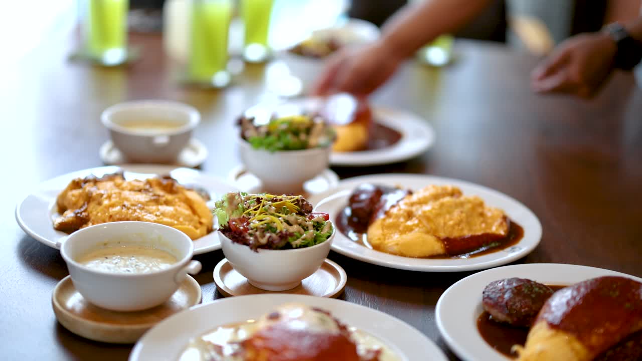 Server places omurice and hamburg steak plates on restaurant table under soft natural lighting