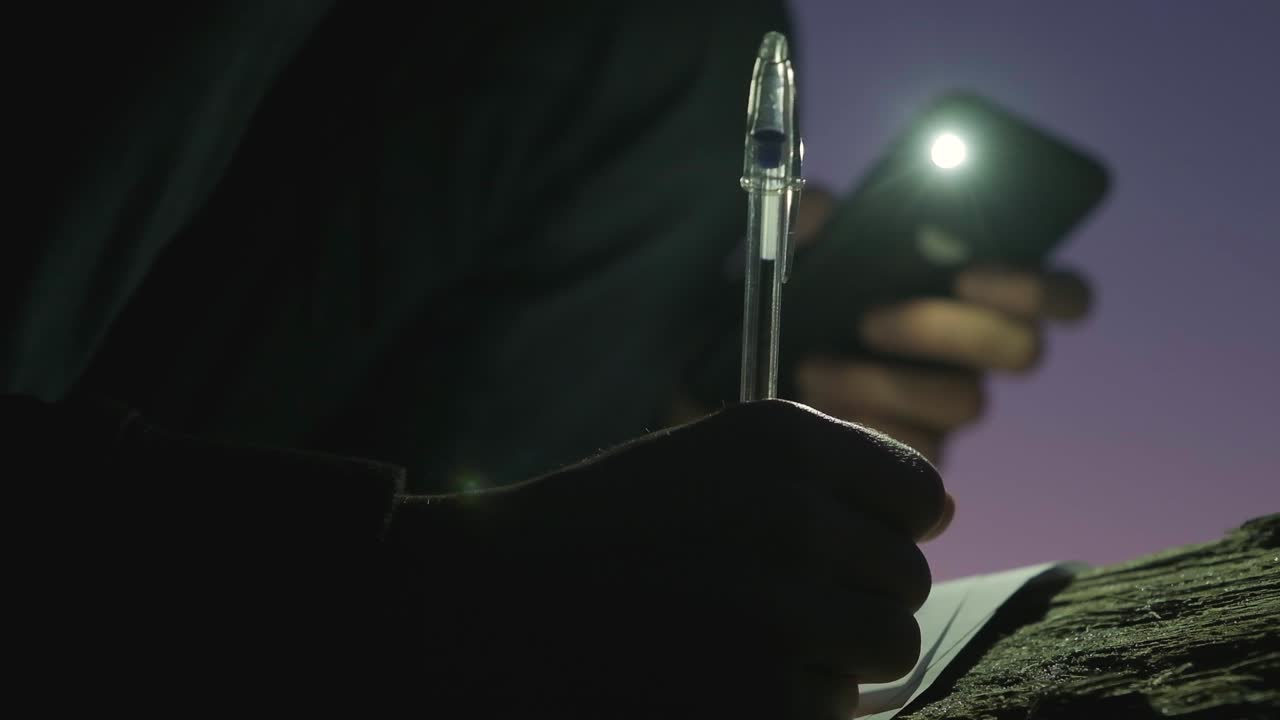 A person writing on paper at night, shining a cellphone torch on the paper.