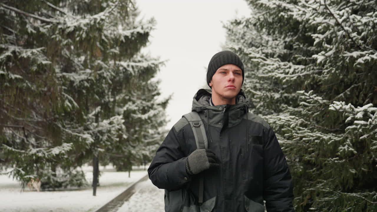 Winter park walkway closeup of pedestrian, young man in black jacket and beanie carrying backpack, steady stride toward camera, snow on evergreen branches, brisk commute mood, crisp cold light