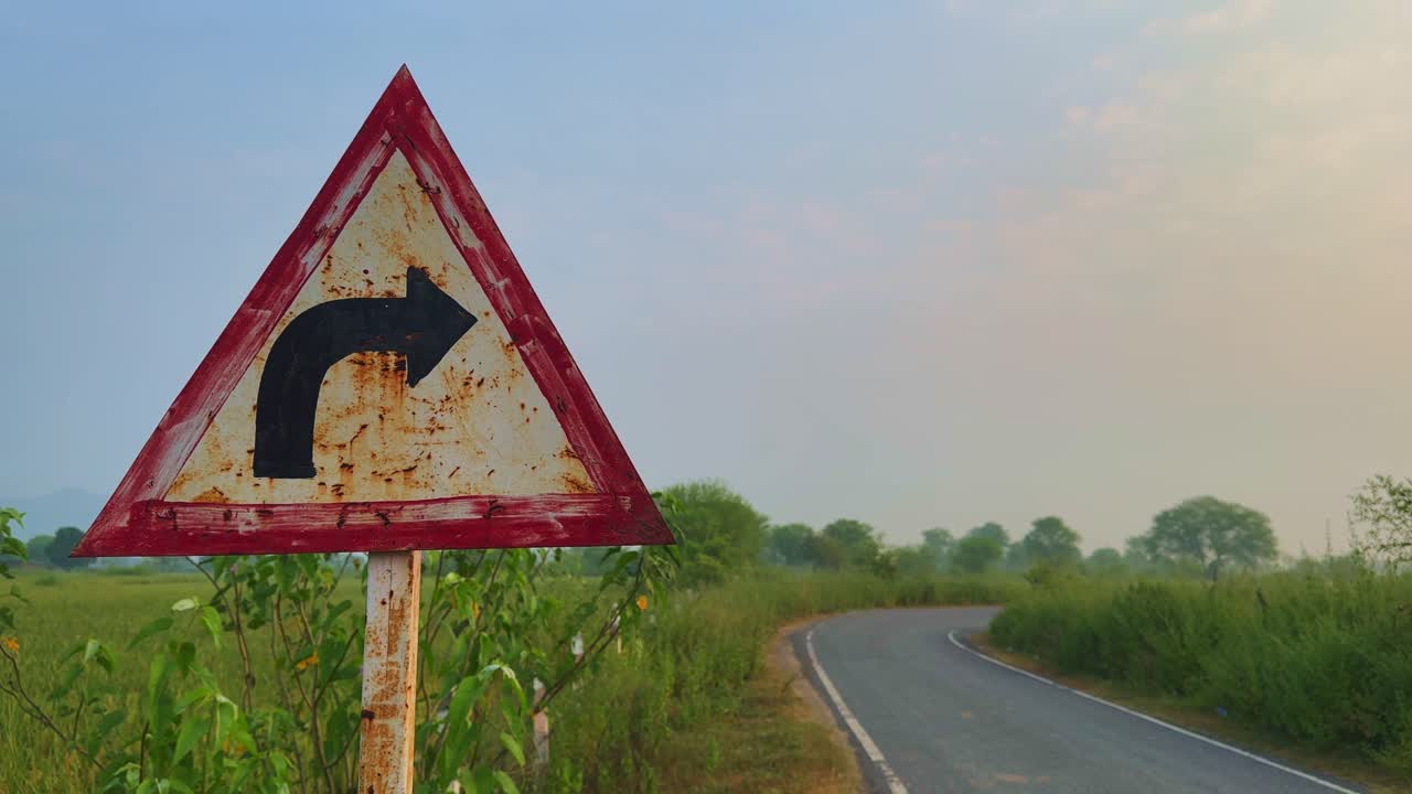A static shot of a weathered right turn road sign on a quiet countryside road during a calm morning, with soft sunlight and greenery surrounding the path