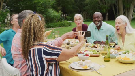 animation of diverse happy senior female and male friends eating lunch in garden, using smartphone