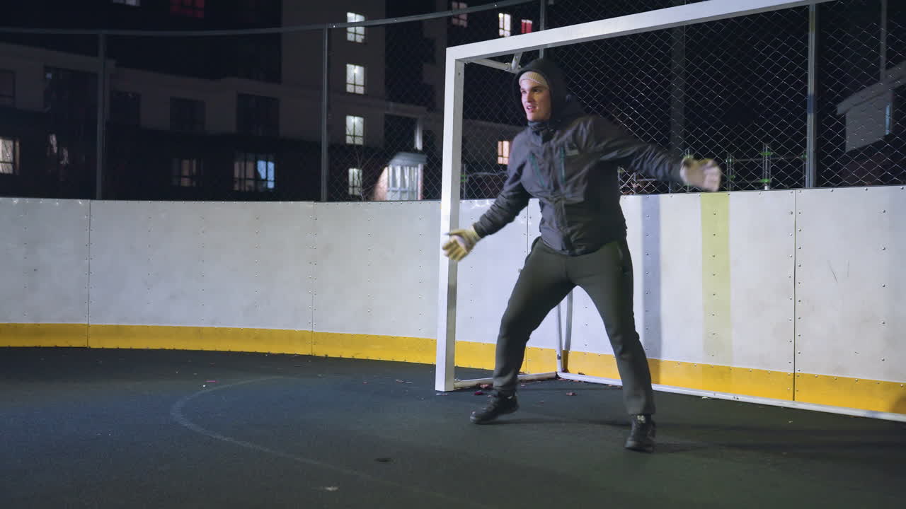 Goal keeper practicing defensive skills near goal post during night training session on outdoor sports court, scene captures athletic preparation with illuminated urban background