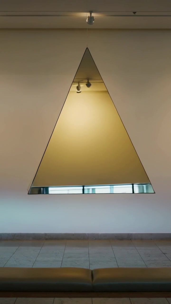 Video verticale: Scultura a piramide specchiata rotante su una panca della galleria, che riflette luci e un cartello.
