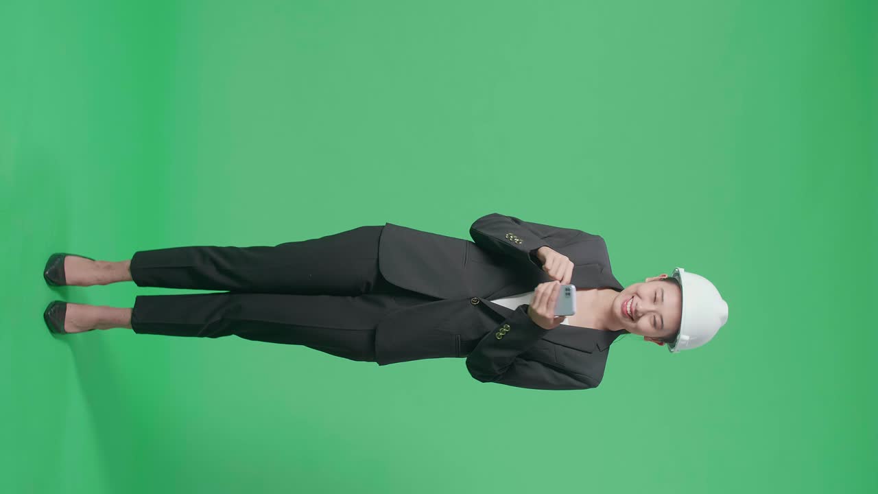 Full Body Of Smiling Asian Female Engineer Using Smartphone And Pointing To The Side In The Green Screen Studio