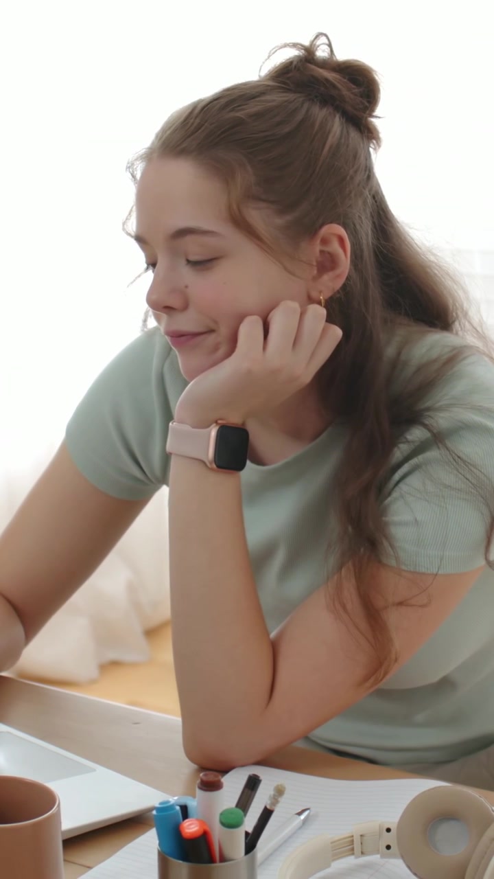 Video verticale: Ragazza adolescente con maglietta verde che studia, guarda il laptop e allunga la mano per prendere una tazza.