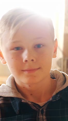 young boy with light blonde hair and blue eyes is smiling warmly, wearing a plaid shirt and hoodie, captured in a softly lit indoor environment with natural light