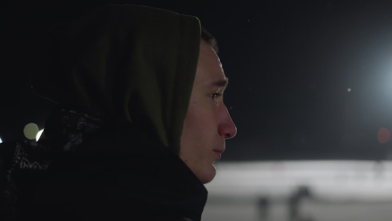 Side view of unhappy boy in hoodie standing outside during night with bright light glowing in distance, capturing mood of isolation and introspection in cold evening atmosphere