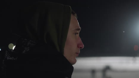 side view of unhappy boy in hoodie standing outside during night with bright light glowing in distance, capturing mood of isolation and introspection in cold evening atmosphere