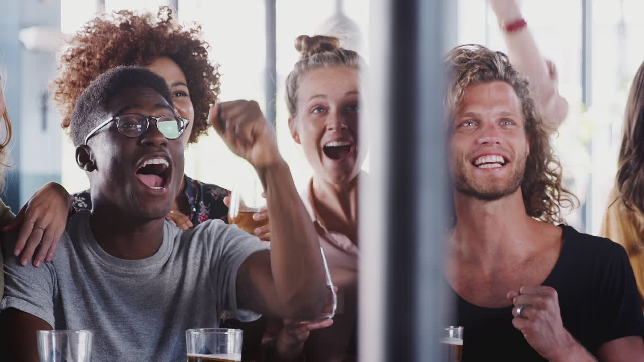 Gruppo di amici e amiche che festeggiano mentre guardano una partita sullo schermo in un bar sportivo