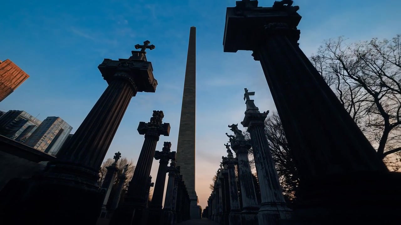Ripresa in tracking con telecamera a bassa angolazione di un obelisco centrato in un viale commemorativo, mentre il crepuscolo cala e le colonne si oscurano.