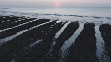 Low-Angle-Drone-shot-flying-over-Bingin-Beach-low-tide-reef-with-sunset-and-human-silhouettes-in-Uluwatu-Bali-Indonesia