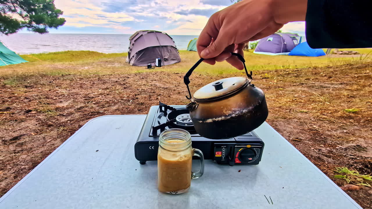 Man pouring hot water from a kettle to make coffee on a portable stove at a campsite with tents by the Baltic Sea - first-person point of view (POV)
