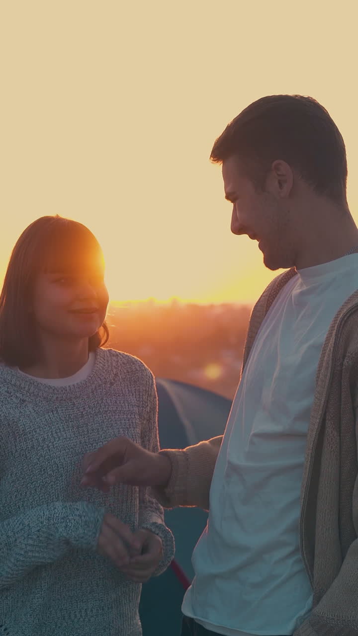lovely young couple joins hands near tent and friends in tourist camp in warm autumn evening against river at sunset
