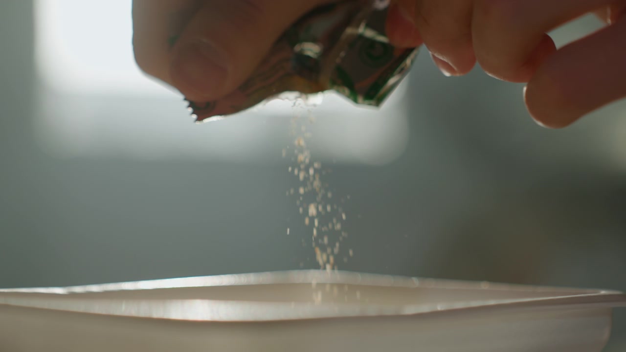 Close-up de mãos polvilhando pó em uma tigela, brilho suave da janela em contraluz, grânulos delicados caindo de um pacote de alumínio rasgado, clima cinematográfico em câmera lenta, preparação tranquila na cozinha e detalhes de textura