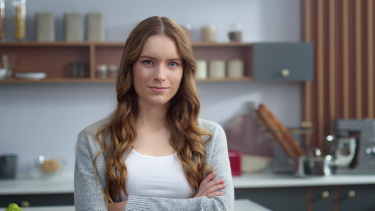 Atraktivní žena se dívá do kamery v kuchyni. Soustředěná dívka se zkříženýma rukama.