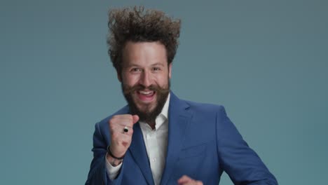 dancing curly hair man with fists up having fun, being dynamic, energy and enjoying, making funny faces, partying, dancing and moving on blue background in studio