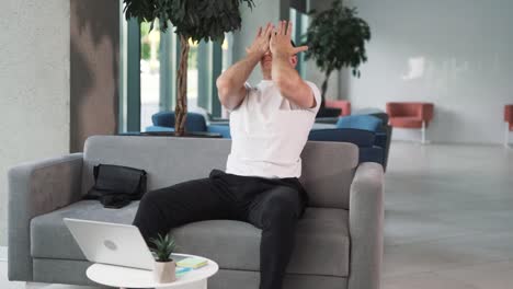 close-up-portrait-of-a-young-man-in-a-white-T-shirt-against-the-backdrop-of-a-business-center,-sitting-on-a-sofa,-passionately-following-a-sports-match-on-his-laptop