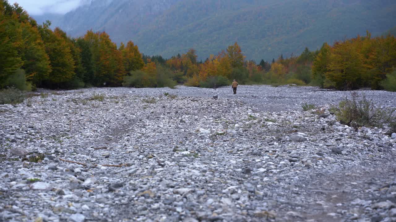 Lonely old man walking on dry riverbed across colorful trees of valley forest at Autumn in Albanian Alps