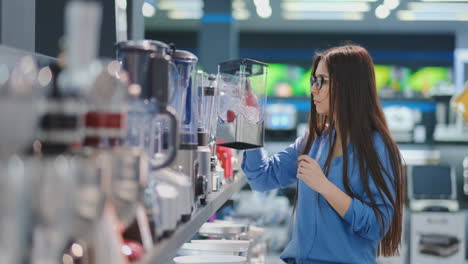 Young-beautiful-woman-in-the-appliances-store-is-choosing-for-its-kitchen-a-blender-looking-and-holding-in-her-hands-various-models.