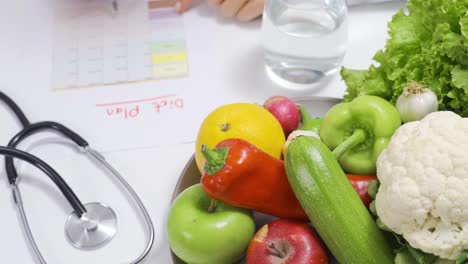 nutricionista mujer escribiendo un plan de dieta en una mesa llena de frutas y verduras.