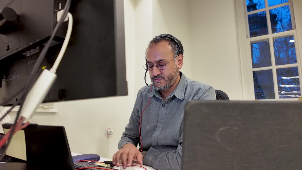 South Asian Man Wearing Headset Working Behind Monitor And Laptop In Office Customer Service Technical Support Call Center Communication Technology
