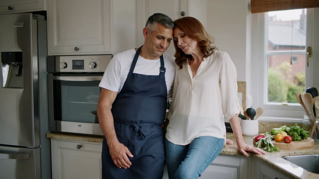 Leaning woman on partner's shoulder, man in navy apron tilting in kitchen, sharing quiet pause