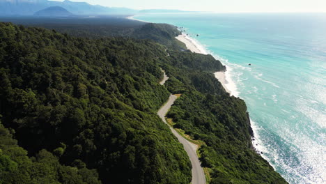 Vista-Aérea-De-Nueva-Zelanda-Del-Clima-Soleado-En-La-Costa-Oeste-Con-Carretera-Asfaltada-Siguiendo-La-Costa-Del-Agua-Azul-Del-Océano-En-Un-Bosque-Verde-Y-Exuberante