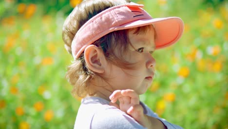 Face-of-Shy-and-sad-child-girl-in-pink-hat-in-the-middle-of-grassy-meadow-with-yellow-flowers---slow-motion