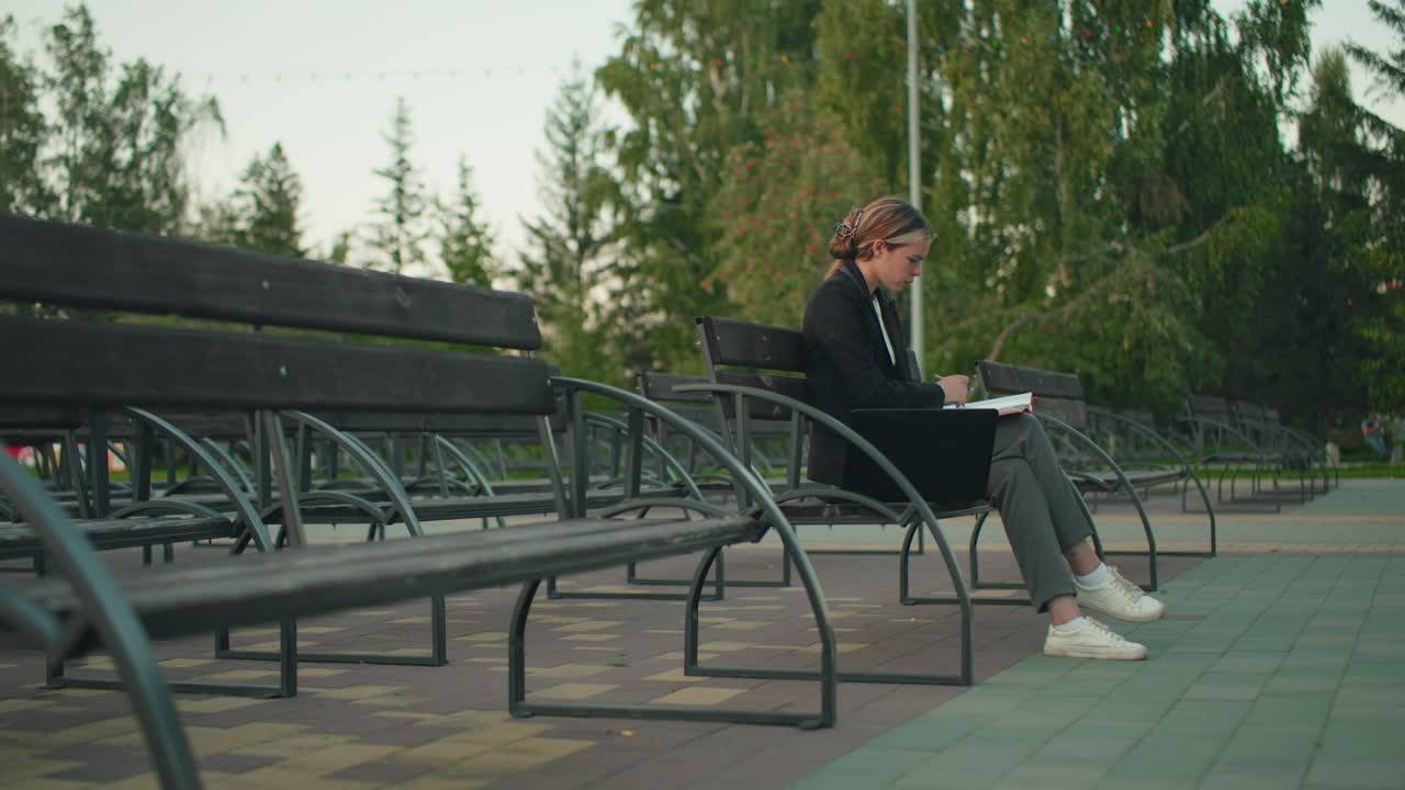 Woman in professional attire seated alone on outdoor bench flipping through red folder with pen, laptop beside her, surrounded by empty benches in quiet park with green trees and paved walkway