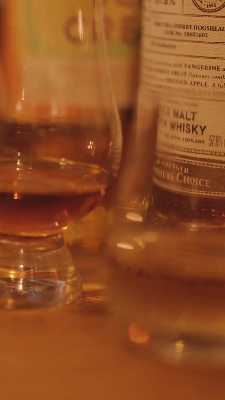 Un'inquadratura a fuoco selettivo di bicchieri e bottiglie di whisky su una superficie di legno, con illuminazione ambientale morbida e calda e movimento minimo della telecamera, creando un'atmosfera accogliente