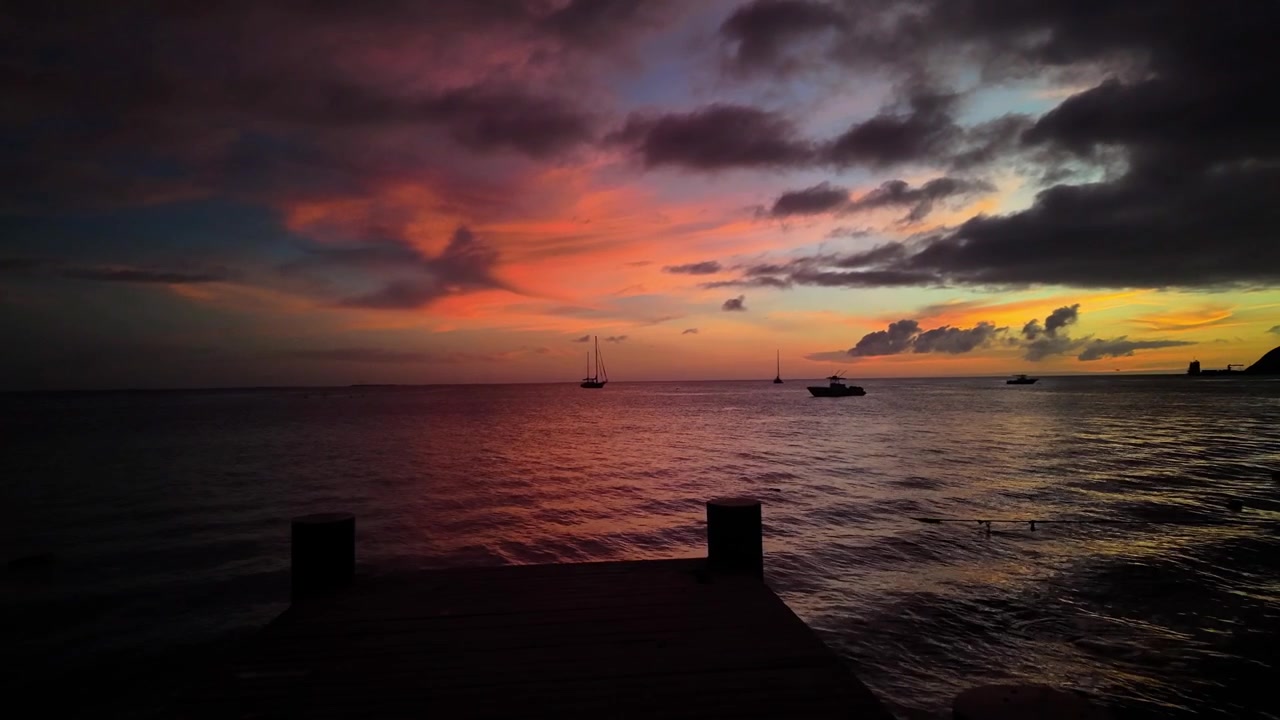 Eine ruhige Weitwinkelansicht eines einsamen Betonstegs, der sich in das ruhige karibische Wasser erstreckt, während eines lebhaften, farbenfrohen Sonnenuntergangs mit Booten als Silhouette.