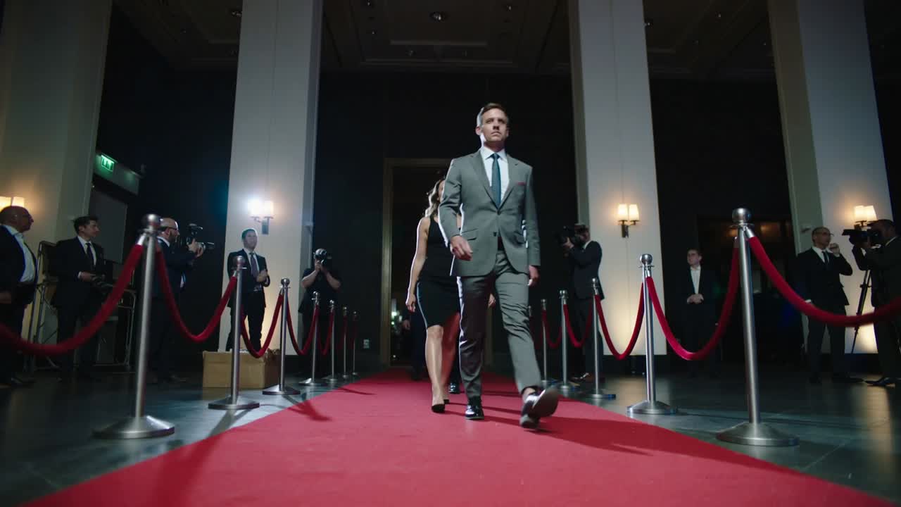 Walking gray-suited man leading group moving along red carpet toward entrance opening, with cameras