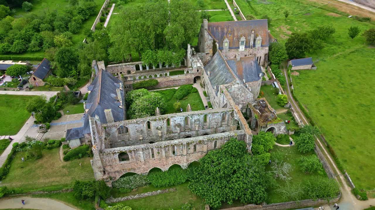 Drone shot pulling back from Beauport Abbey ruins, revealing gardens, paths, and the low tide beach - Frabce