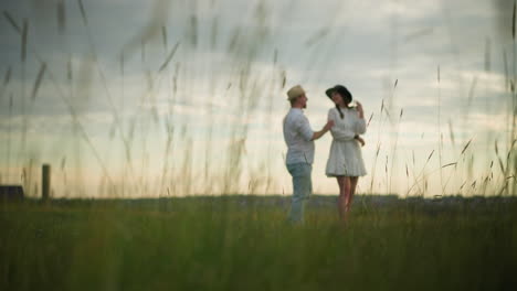 a tranquil scene where a man in a hat, white shirt, and jeans walks toward a woman in a white dress standing on a grassy hill at sunset