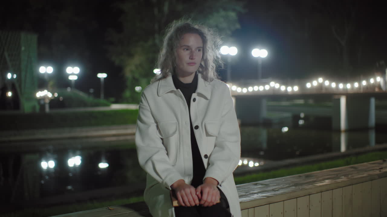 Woman in white coat sits by calm water at night, enjoying cool evening ambiance, lights reflecting gently, capturing peaceful moment of quiet solitude in open urban setting, hand resting on book