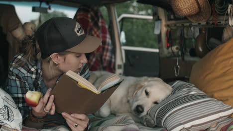 woman eating apple and reading book while lying with dog in van