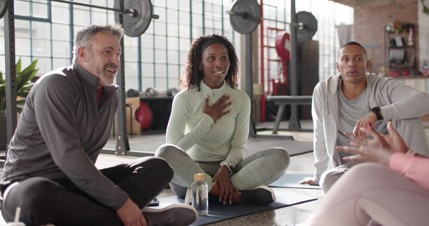 Gruppo fitness eterogeneo seduto sui tappetini in palestra, che esprime gratitudine all'istruttrice in leggings rosa.