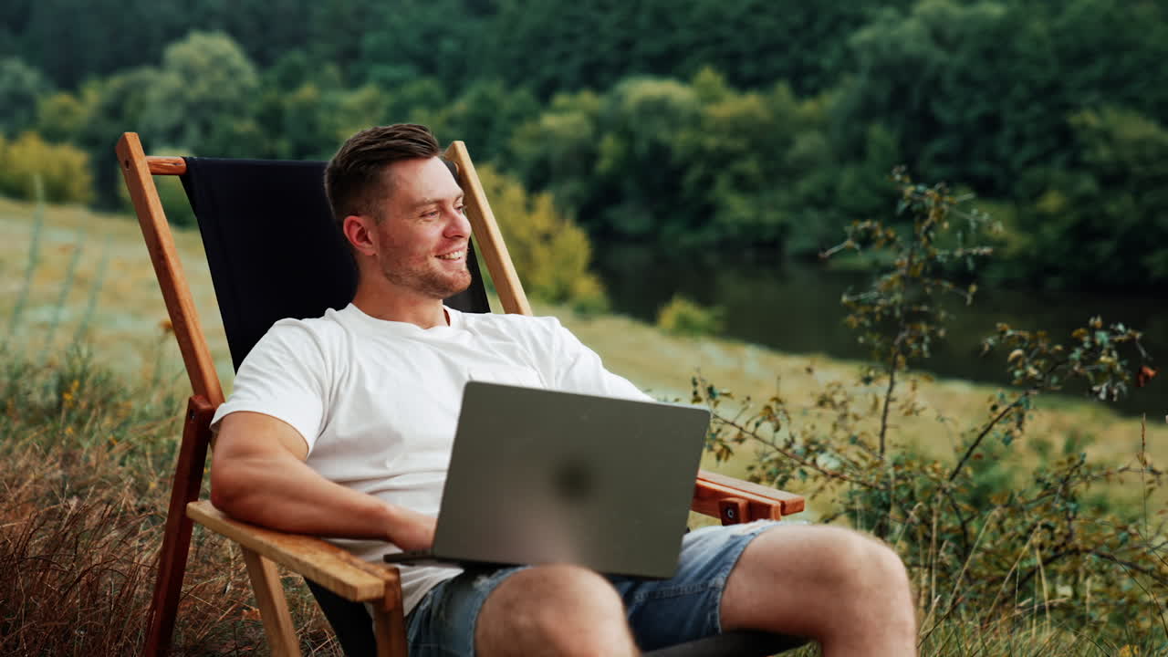 Thoughtful Caucasian man sits holding a laptop smiling dreamily. Freelancer working remote in a comfortable natural surroundings.