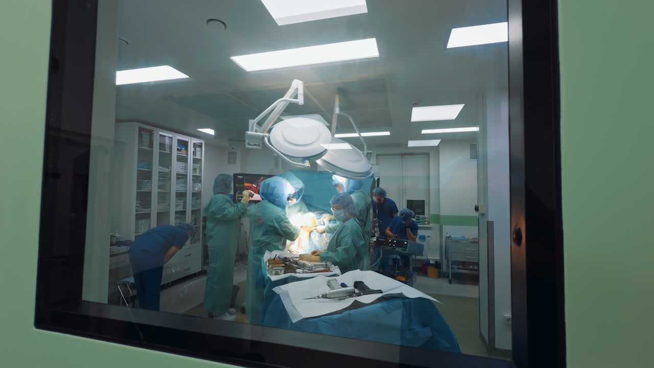 Kyiv, Ukraine, 2 August 2025: Operating room activity viewed through a small, square window in a green wall. The distant perspective shows surgeons and medical equipment under intense illumination and dome lights