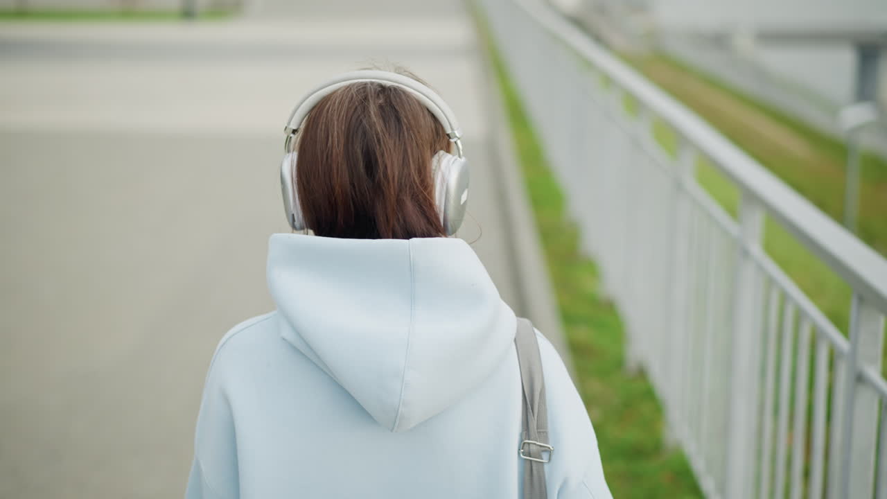 Back view of young girl in sweater and headphones walking, looking towards blurred iron rail in background, calm, relaxed outdoor scene with focus on her and surrounding environment