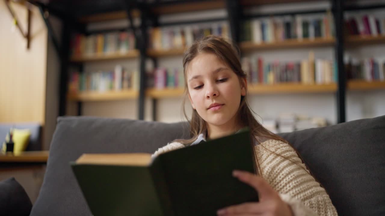 Teenage girl reading a book on a sofa in a library-like interior