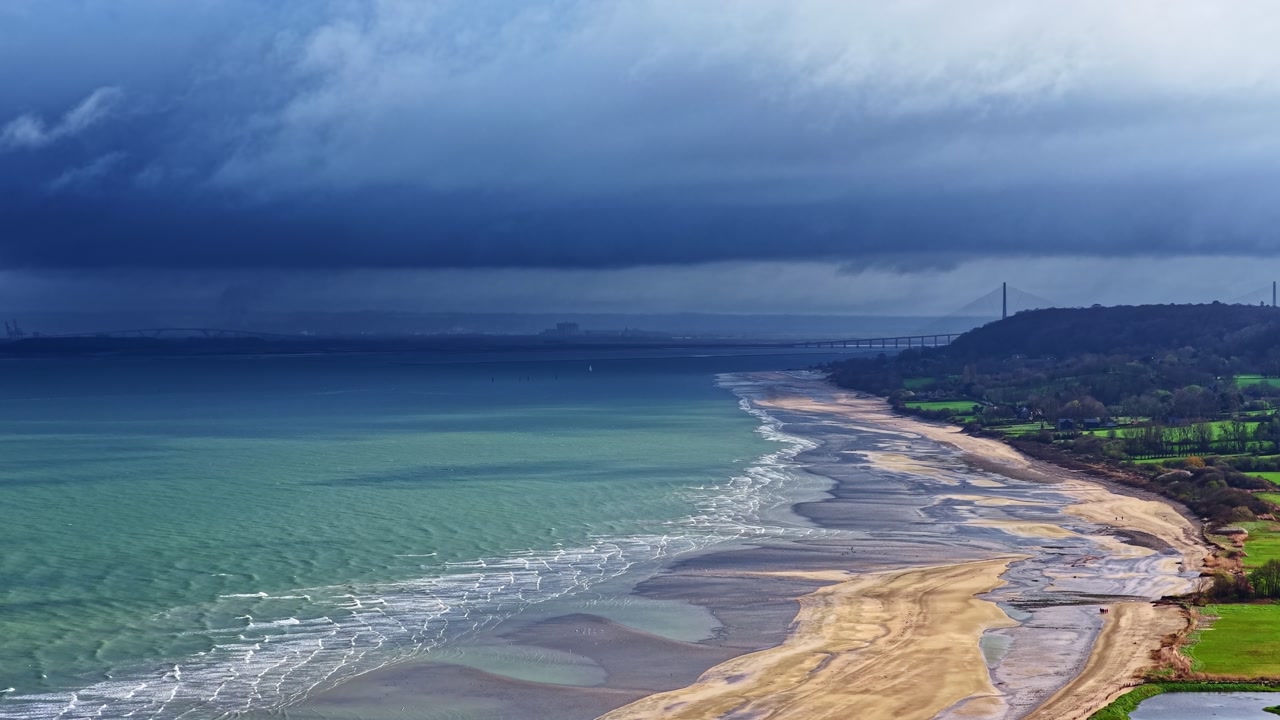 Aerial view of a storm front rolling over the Normandy coast, with turquoise sea, wide sandy shoreline, and dramatic clouds creating a moody cinematic seascape.