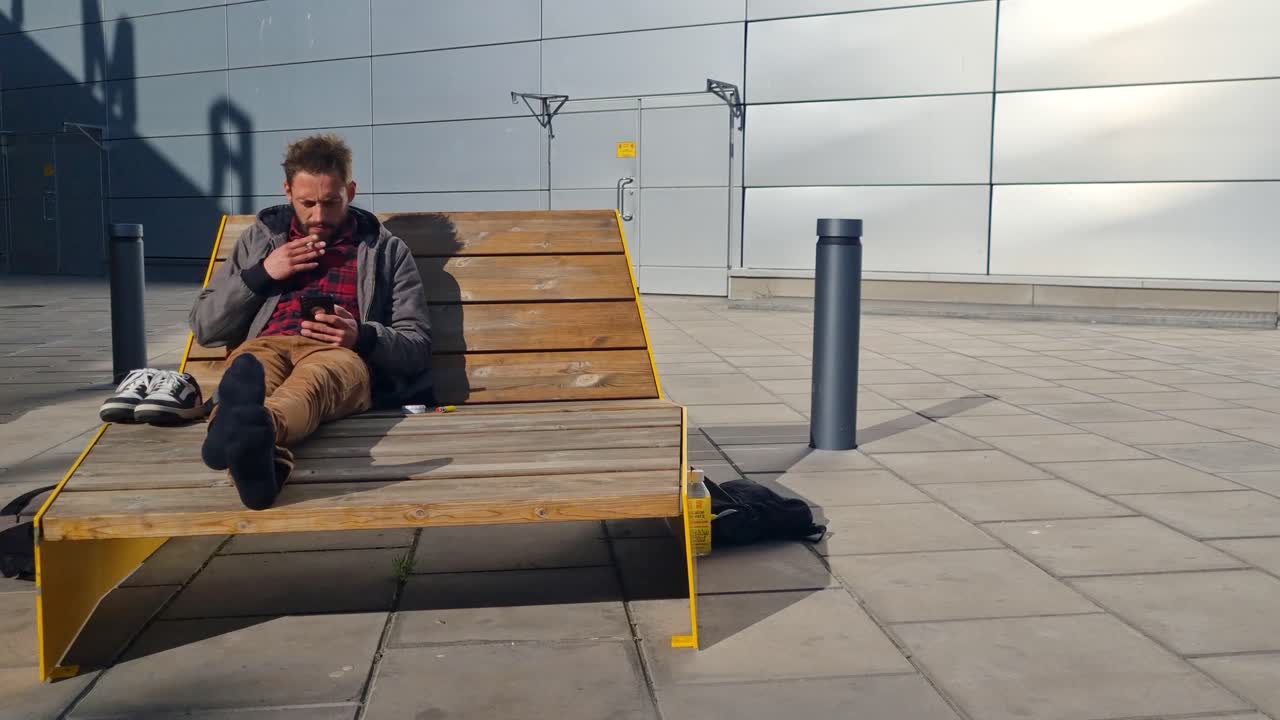 A man sits relaxed on a wooden bench at Arlanda Airport, casually smoking and scrolling on his smartphone—an unhurried moment in transit