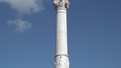 Lisbon,-Rossio-Square,-Pedro-4th's-statue's-tilt-up,-detailed-footage-of-the-white-sculptures-at-the-base-and-the-bronze-Pedro-at-the-top-on-a-sunny-day-with-blue-skies-and-white-clouds-at-the-end