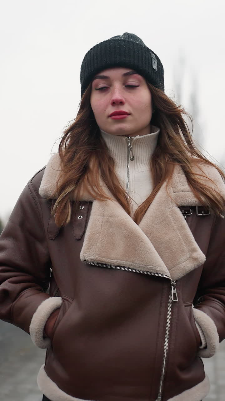 Portrait shot of lady in black cap brown shearling jacket walking slowly with hands in pockets on wide stone pathway through quiet park lined with pine trees modern buildings and cloudy autumn sky